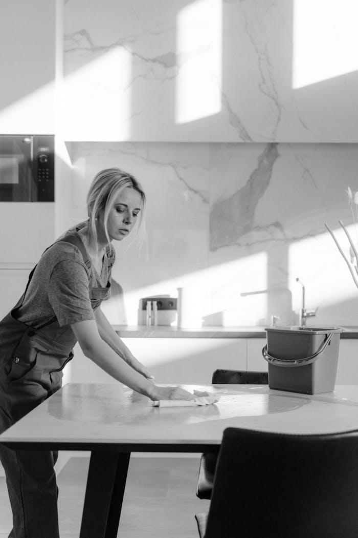 A female cleaner sanitizing a kitchen table with natural light casting shadows.